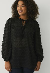 Zwarte blouse van een gestructureerde, gemusterde stof. Heeft een ronde halslijn, strikdetail en volumineuze mouwen. Zacht en lichtgewicht materiaal.