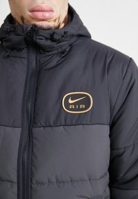 Czarna kurtka puchowa z wysokim kołnierzem, z złotym logo Nike AIR na klatce piersiowej i zamkami błyskawicznymi z przodu.