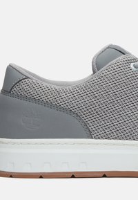 Zapatilla de malla gris con un panel trasero gris suave, media suela blanca y suela de goma marrón. Presenta un logo sutilmente grabado.