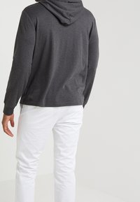 Grijze hoodie met lange mouwen; zachte textuur en een relaxte pasvorm. Draagt met witte broek, wat een casual outfit laat zien.