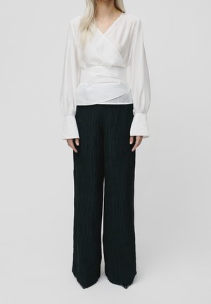 Kobieta ubrana w białą bluzkę z kopertowym zapięciem i dużymi mankietami oraz szerokie, czarne spodnie z teksturą, stojąca na tle jednolitego tła.