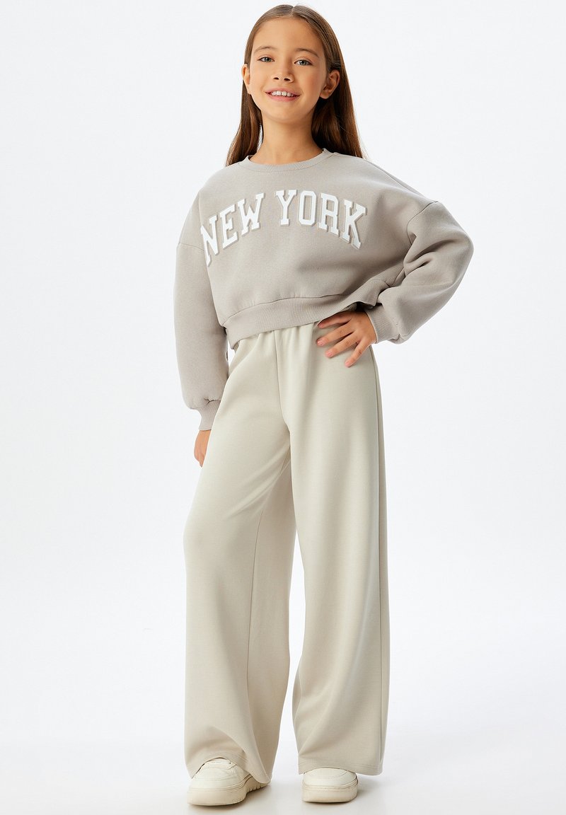 Grijze sweatshirt met de tekst "NEW YORK", gecombineerd met losse crèmekleurige broeken. Model draagt witte sneakers. Soepele stof, relaxte pasvorm.