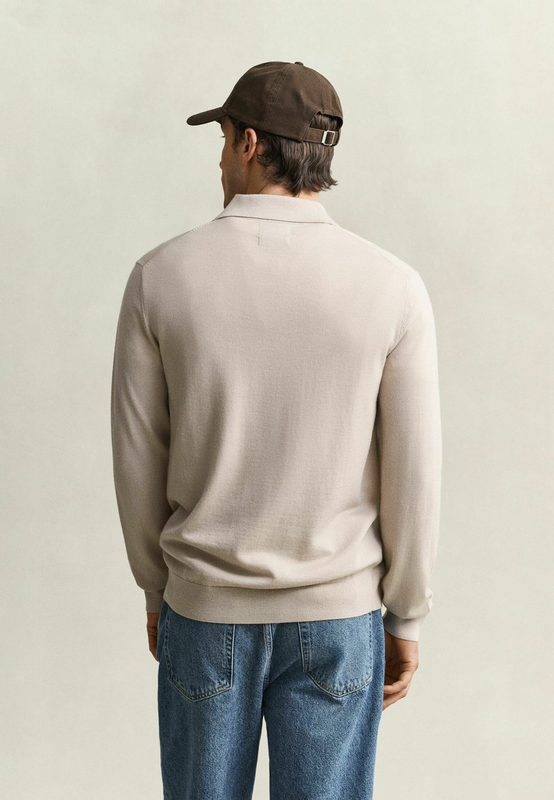 Man wearing a beige long-sleeve shirt, blue jeans, and a dark brown baseball cap facing away against a plain background.