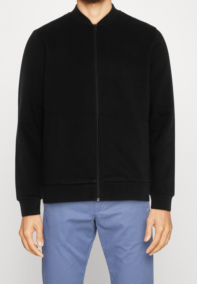 Czarna bluza rozpinana na zamek z zaokrąglonym kołnierzem, wykonana z miękkiej tkaniny; posiada ściągacze przy mankietach i u dołu, minimalistyczny design.