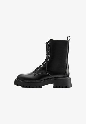 Botte de combat noire en cuir à lacets avec semelle en caoutchouc épaisse et languette à l’arrière, vue de côté sur fond blanc.