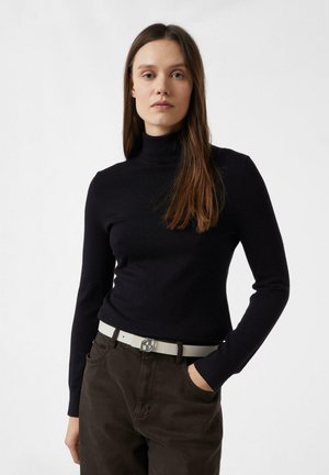 Kobieta z długimi brązowymi włosami, ubrana w czarny sweter z golfem, ciemne brązowe spodnie i biały pasek, stojąca z jedną ręką w kieszeni.