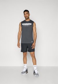 Siva brezrokavni športni top z napisom "RAIDERS Football" v belem, v kombinaciji s temnimi kratkimi hlačami; črno-bele športne čevlje in bele nogavice.