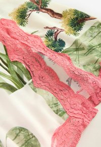Stoff mit Floralprint, der grüne Blätter und Bäume zeigt, mit einem auffälligen rosa Spitzenbesatz, der das Design überlagert und einen strukturierten Kontrast schafft.