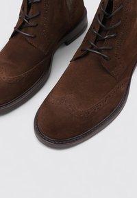 Botas brogue de ante marrón con costuras decorativas y acentos perforados, con puntera redondeada y suela plana de cuero.