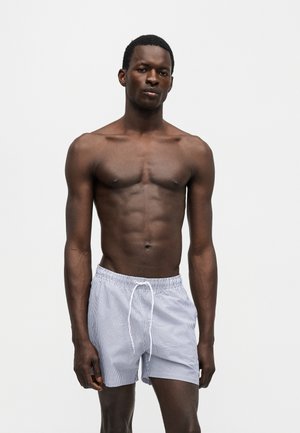 Homme à la peau foncée portant un short de bain rayé bleu clair et blanc avec un cordon, debout devant un fond blanc uni.