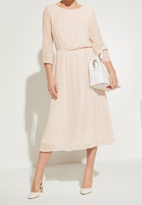 Pfirsichfarbendes Kleid mit Polka-Dots, dreiviertelärmeln, tailliertem Bund und einem fließenden Saum, kombiniert mit einer weißen Handtasche und hohen Schuhen.