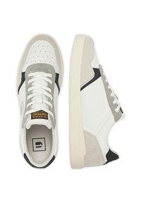 Baltas un pelēkas krāsas sporta apavi ar zamšādas un ādas virsmu, baltām plakankājām šņorēm, perforētu purngali un kontrastējošiem melniem akcentiem.