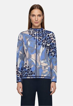 Vrouw die naar voren kijkt, draagt een blouse met lange mouwen met blauwe en beige bloemen- en luipaardprint en een donkere broek.
