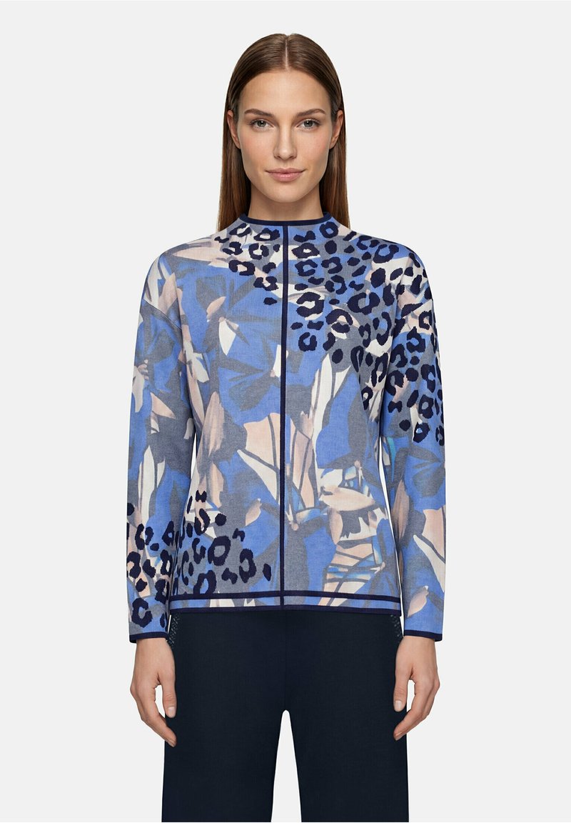 Femme de face portant une chemise à manches longues avec des motifs floraux et léopard bleus et beige et un pantalon foncé.