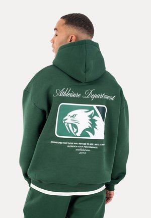 Mann trägt einen dunkelgrünen Hoodie mit dem Aufdruck "Athleisure Department" und einem brüllenden Wildkatzen-Logo auf dem Rücken.
