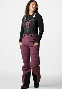 Een vrouw in paarse buitenbibs met zwarte patches en een zwarte lange mouwtop, staand met haar handen in haar zakken en glimlachend.