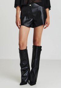 Bermuda in pelle nera con vita alta, cuciture a contrasto e tasche frontali, abbinate a stivali in pelle nera al ginocchio con cerniere.