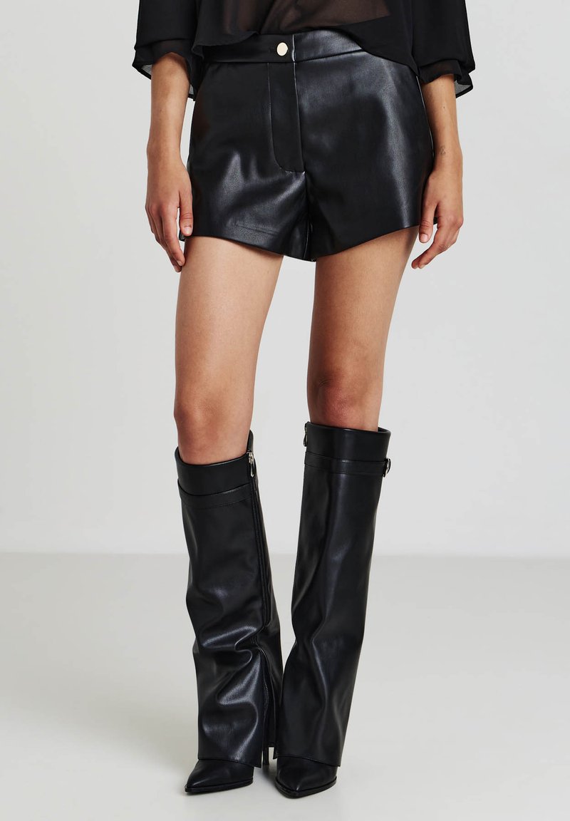 Bermuda in pelle nera con vita alta, cuciture a contrasto e tasche frontali, abbinate a stivali in pelle nera al ginocchio con cerniere.