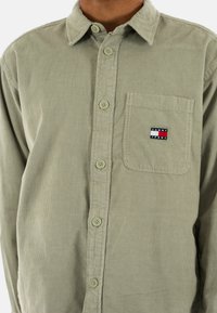 Olive green corduroy shirt featuring a button-up front, collar, and a chest pocket with a "Tommy Jeans" label. Textured fabric with vertical ridges.