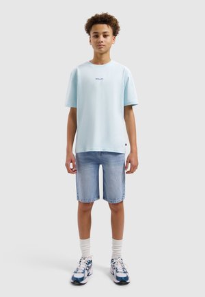 Tienerjongen die recht vooruit kijkt, gekleed in een lichtblauw T-shirt, spijkershorts, witte sokken en blauw-witte sneakers tegen een effen achtergrond.