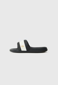Sorte svarte sandaler har en bred stropp med hvit aksent og en gull logo. Sålen er teksturert for grep og er laget av gummimateriale.