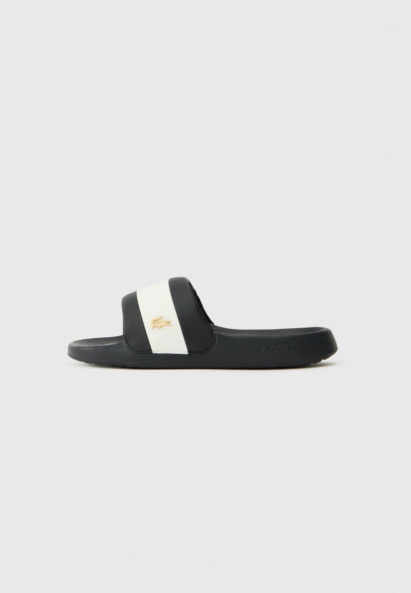 Sorte svarte sandaler har en bred stropp med hvit aksent og en gull logo. Sålen er teksturert for grep og er laget av gummimateriale.