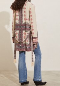 Mujer vestida con un cardigan beige de patrón, con cintas largas, jeans azules y botas de tacón negras, de pie contra una pared lisa.