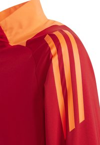 Rotes Sportshirt mit Stehkragen und orangenen drei Streifen akzenten an den Schultern. Glattes, leichtes Material.