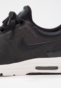 Musta urheilukengä, jossa on teksturoitu päällinen, hienovarainen Nike-kaarre, pehmustettu nilkka-aukko ja kontrastoiva valkoinen välikerros.