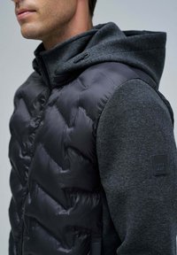 Gilet en doudoune sans manches noir avec un design matelassé, associé à un sweatshirt à capuche gris foncé. Présente une étiquette de logo sur la manche.