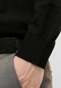 Juodas megztinis su elastingu audiniu, rankovės kraštas su raštu. Rankos matomos, laikančios audinį, fone juodas diržas. Apačioje gradientinės kelnės.