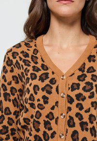 Cardigan en imprimé léopard dans des tons beige et noir, avec un col en V, des poignets côtelés et des boutons marron à l'avant. Texture tricot douce.