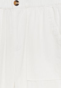 Gros plan sur un short en tissu blanc avec une taille élastique, une grande poche avant plaquée et un unique bouton marron marbré en haut.