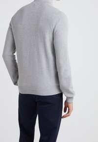 Grått stickat tröja med hög krage, ribbade ärmslut och fåll. Tyget har ett strukturerat mönster och bärs över mörkblå byxor.