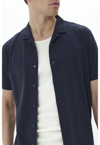 Marinblå kortärmad knapplös skjorta i mjukt tyg, med en platt krage och sex knäppningar. Buren över en vit ribbad linne.