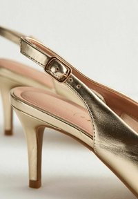 Tacones altos slingback metálicos dorados con correas ajustables y tacones stilettos bajos sobre una superficie blanca.