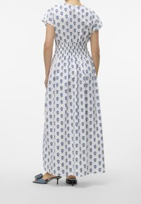 Weißes Maxikleid mit blauem Totenkopf-Print, tailliert und mit kurzen Ärmeln. Kombiniert mit blauen, stoffbezogenen Absatzsandalen.