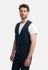 Chaleco azul marino de tejido texturizado, con escote en V, cinco botones y un bolsillo en el pecho. Lleva puesto una camiseta blanca.