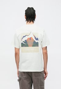 Valge puuvillane T-särk, millel on seljal mägede ja päikeseloojanguga graafiline trükk. Lahtine lõige lühikeste varrukatega, kantud hallide pükstega.