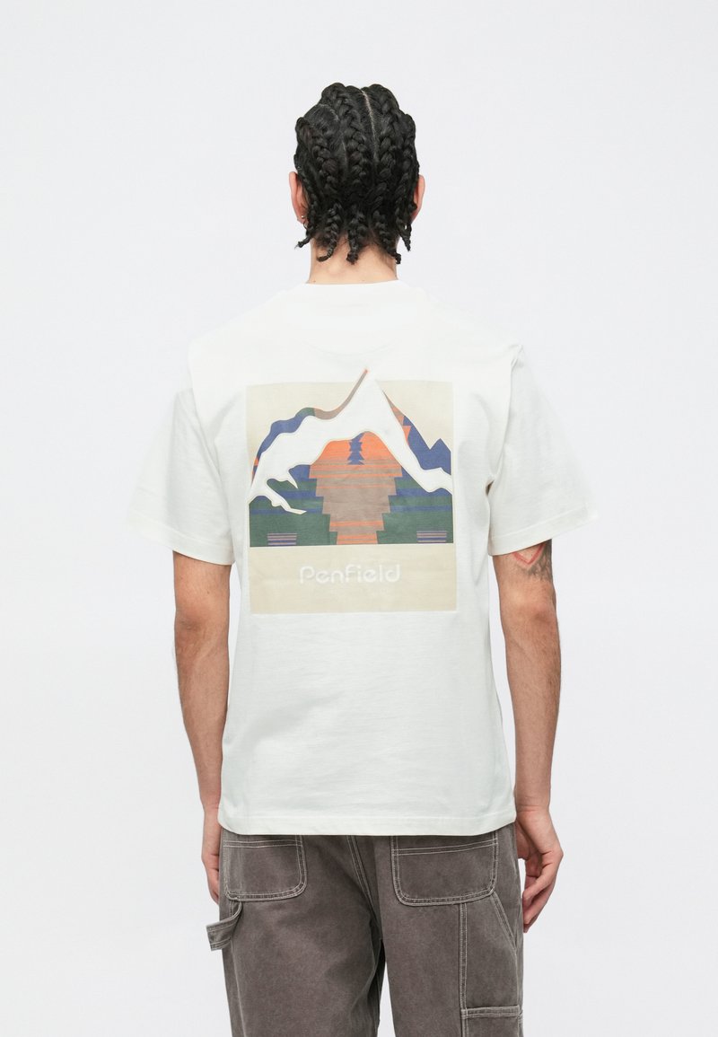 Valge puuvillane T-särk, millel on seljal mägede ja päikeseloojanguga graafiline trükk. Lahtine lõige lühikeste varrukatega, kantud hallide pükstega.