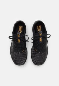 Zapatillas deportivas negras con parte superior de malla, patrón texturizado, puntera redonda y cordones negros. Presenta un logo naranja y etiquetas en el interior.