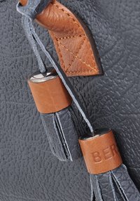 Grijze leren tas met textuur, voorzien van bruine leren accenten en zilverkleurige hardware, die decoratieve franjes en gestikte details heeft.
