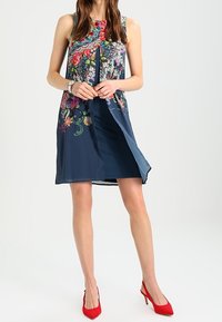 Mekko, jossa on merivoimien väri ja kukkakuosi, hihaton malli ja halkio edessä. Yhdistetty punaisiin korkokenkiin, jotka näyttävät sileiltä ja värikkäilt�ä.