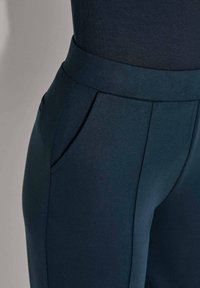 Close-up van een persoon die donkerblauwe, fitted broeken met een brede tailleband en zijzakken draagt, tegen een effen achtergrond.