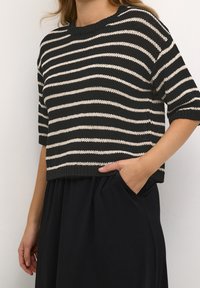 Maglietta a righe nera e crema in maglia con maniche corte e scollo a giro, abbinata a una gonna nera, mani nelle tasche.