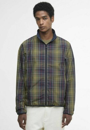 Man met kort krullend haar die een jas met rits draagt met een groen, zwart en rood geruit patroon en witte broek tegen een witte achtergrond.