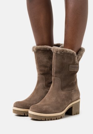 Hnědé semišové kozačky po lýtka s kožešinovou výstelkou, masivními podpatky a robustní podrážkou, nošené na tmavé kůži proti bílému pozadí.