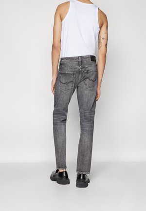 Man draagt vervaagde zwarte slim-fit spijkerbroek, wit mouwloos shirt, zwarte schoenen, staat met de rug naar de camera tegen een effen achtergrond.