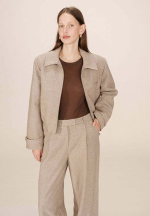 Veste courte en mélange de laine beige avec un col et deux poches avant, associée à un haut ajusté marron et un pantalon taille haute assorti.