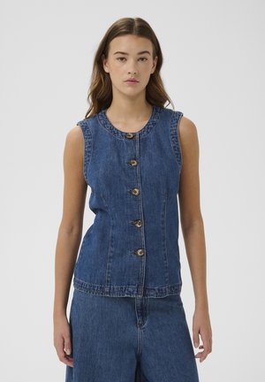 Junge Frau trägt ärmelloses, blaues Jeans-Top mit Knöpfen und passende blaue Jeanshose, steht vor einem schlichten hellen Hintergrund.